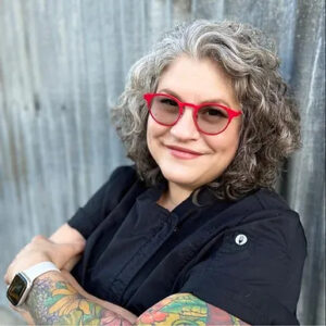Chef Cristina Topham stands crossed armed smiling at camera with chef suit on and red glasses.