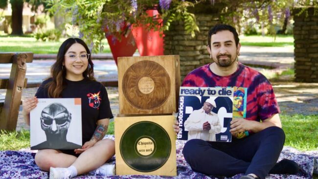 Cera Cera open turntables DJs sitting on a blanket outside holding various records of music.