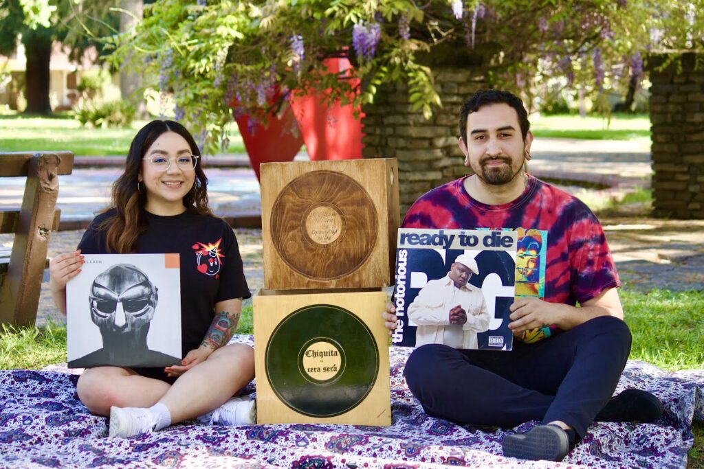 Cera Cera open turntables DJs sitting on a blanket outside holding various records of music.